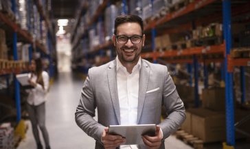 Cheerful and successful middle aged caucasian manager businessman holding tablet computer in large warehouse organizing distribution.
